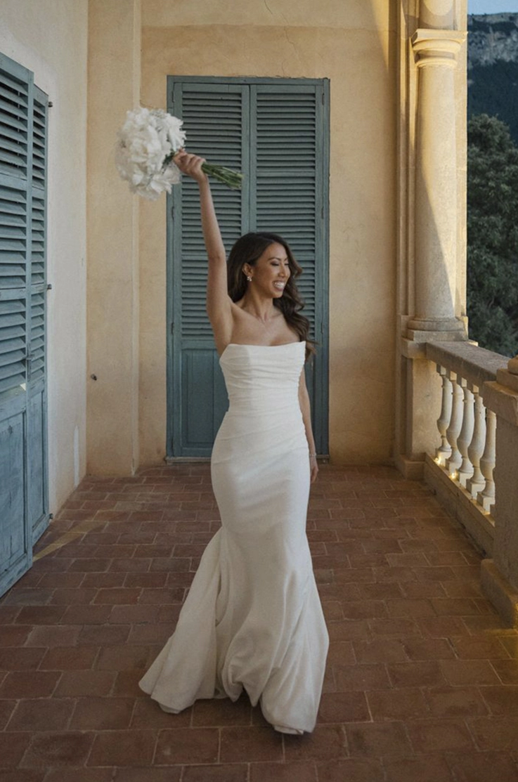 Bride holds a bouquet in the air, in a crepe fit and flare gown with a draped bodice, on a stone terrace 