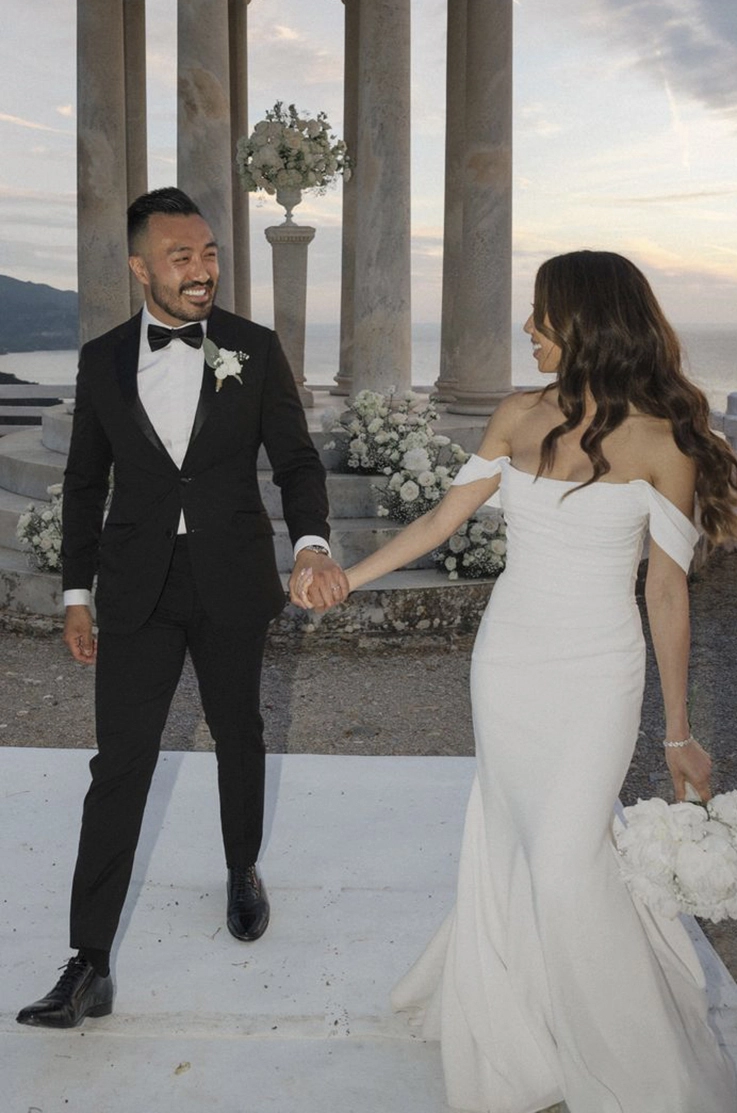 Bride in an off-shoulder crepe fitted gown with draped sleeves, is holding a white bouquet and smiling at groom. 