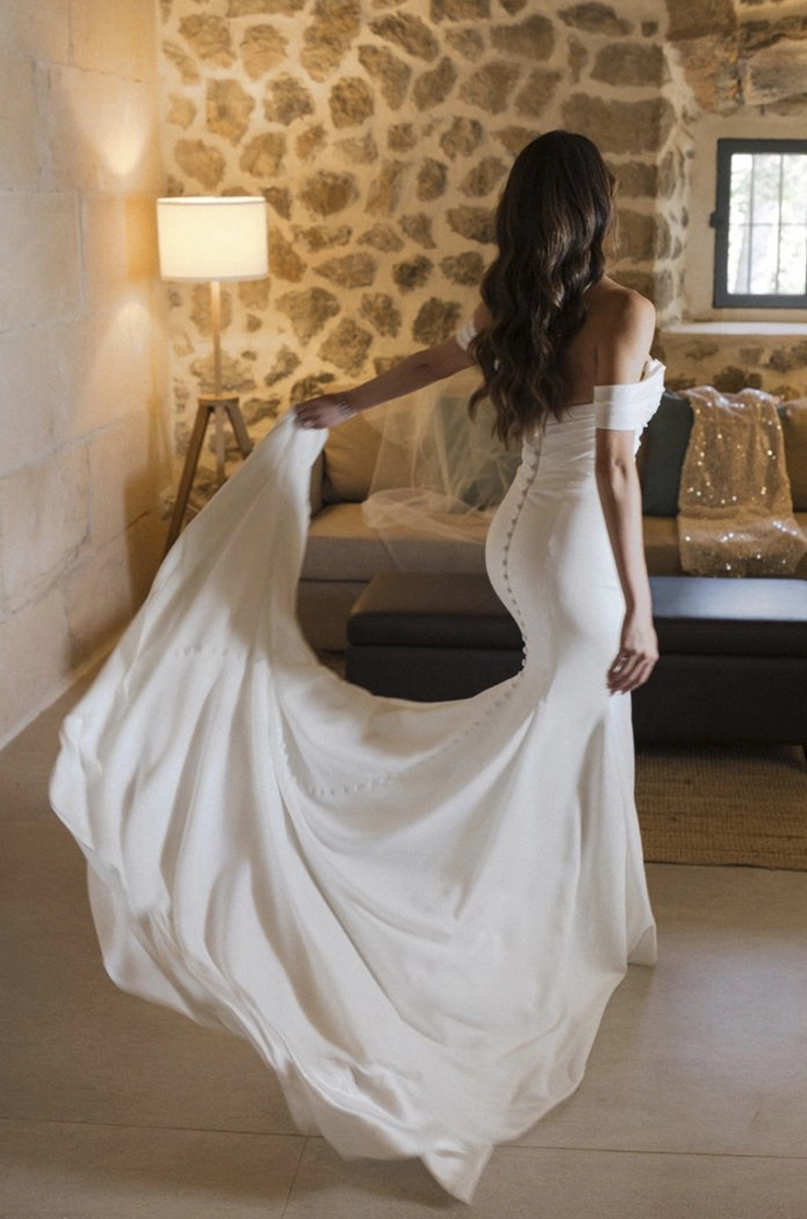 Bride poses and holds her train, in a crepe fit and flare gown with sleeves, draping and a buttoned back by a stone wall. 