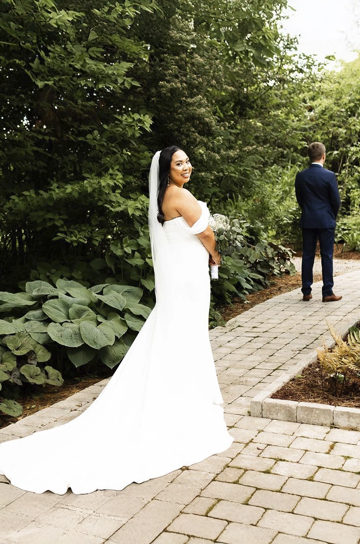 Bride looks back in a crepe fit and flare gown with a veil and draped sleeves, on a stone garden path in front of her groom. 