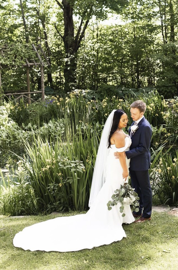 Groom embraces bride in a draped crepe bodice gown with fit and flare skirt and a veil, holding a bouquet next to tall grass.