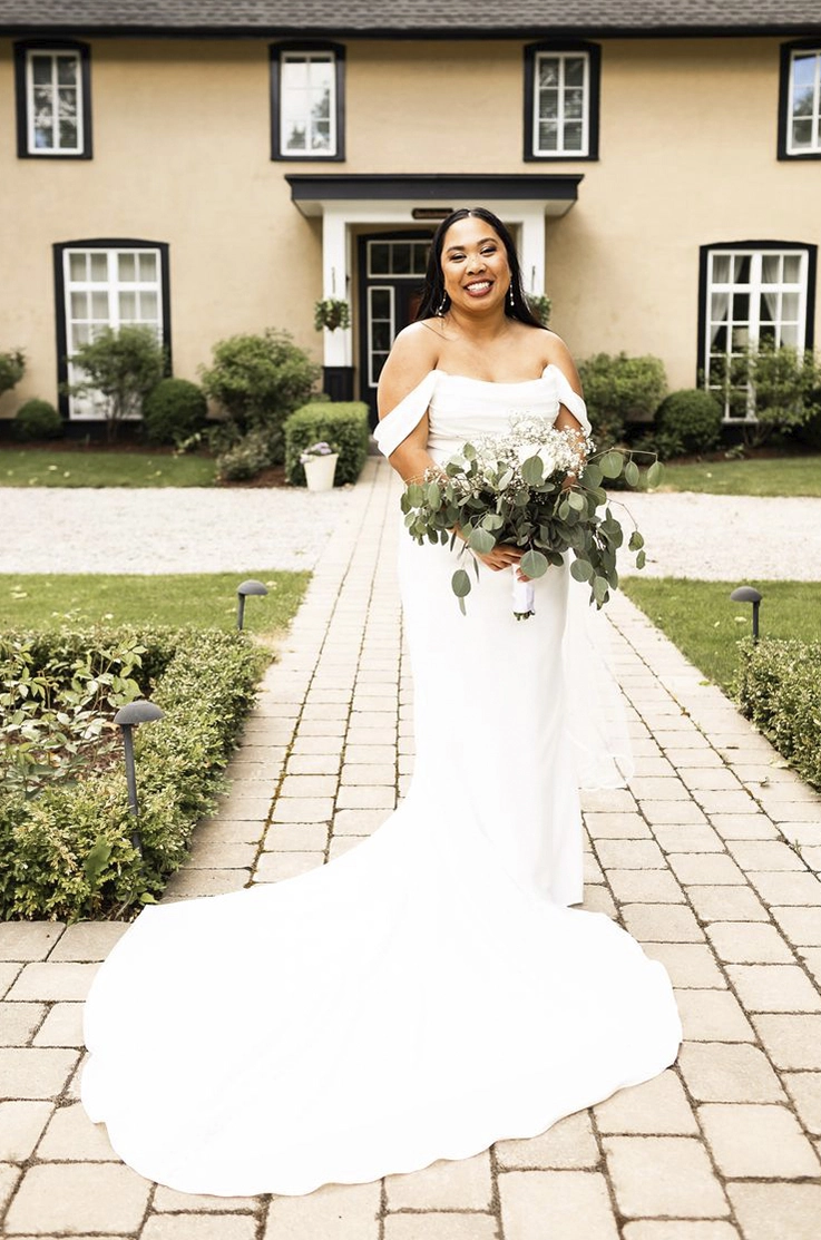 Bride smiles in an off-shoulder fitted crepe gown with a draped bodice and sleeves, on a stone path leading to the venue. 