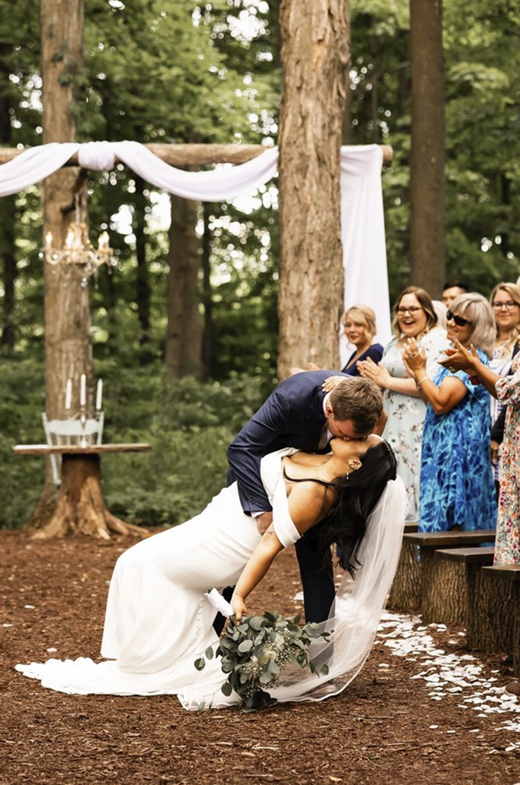 Groom dips his bride in an off-shoulder fit and flare gown with buttons and a veil, and they kiss at a altar in the forest.