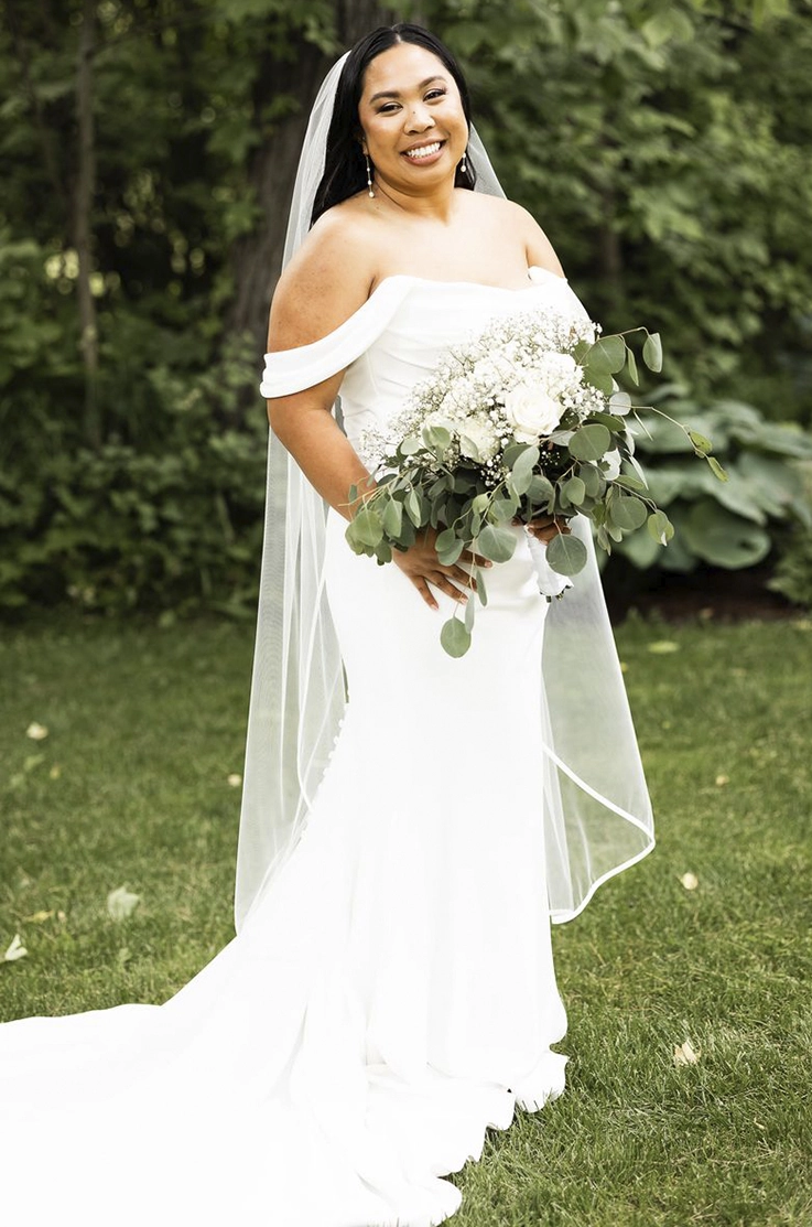 Bride in a draped bodice gown with a fit and flare skirt with buttons and a veil, holds a bouquet on a green lawn. 