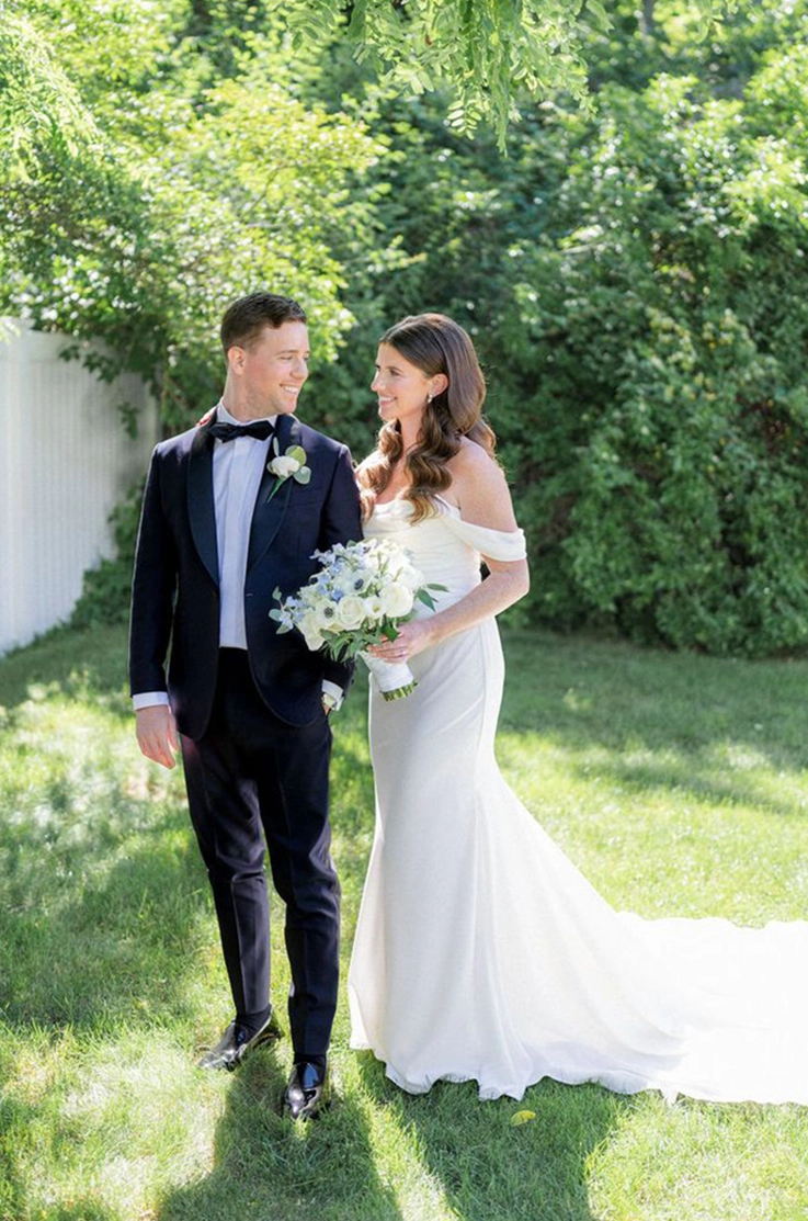 Bride in an off-shoulder fitted gown with a draped bodice and sleeves, poses next to her groom in a sunny backyard. 