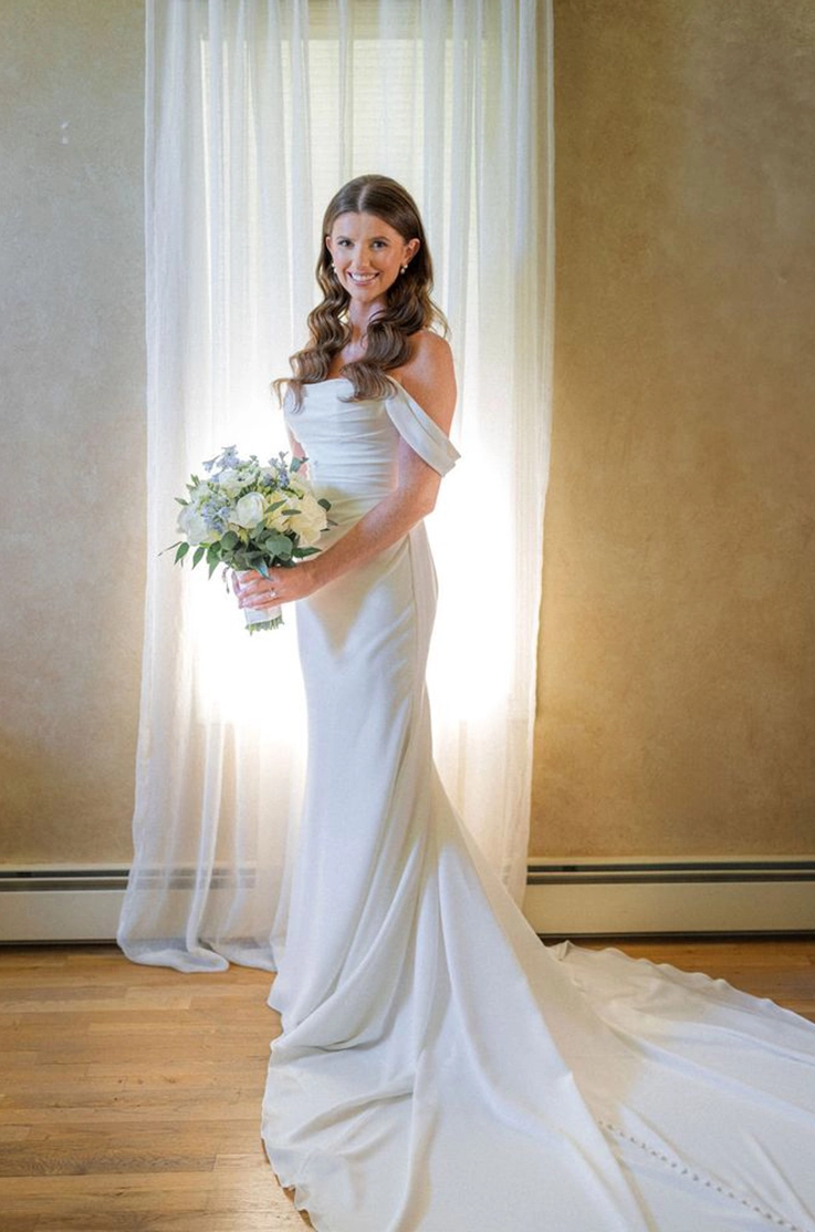 Bride poses by a softly lit window in a draped fit and flare wedding dress with off-shoulder sleeves, holding a bouquet.