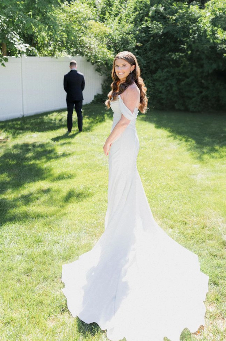 Bride looks over her shoulder in a draped bodice gown with fit and flare skirt and off-shoulder sleeves on a sunny lawn. 