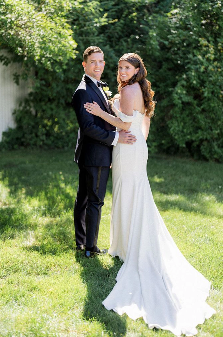 Groom embraces bride in a fit and flare gown with a buttoned back and sleeves, standing on a green lawn.