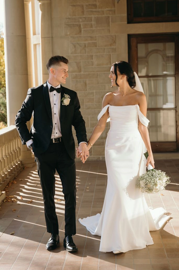 Bride in an off-shoulder fit and flare crepe gown with draping, and a veil, holds hand with groom on a stone terrace.