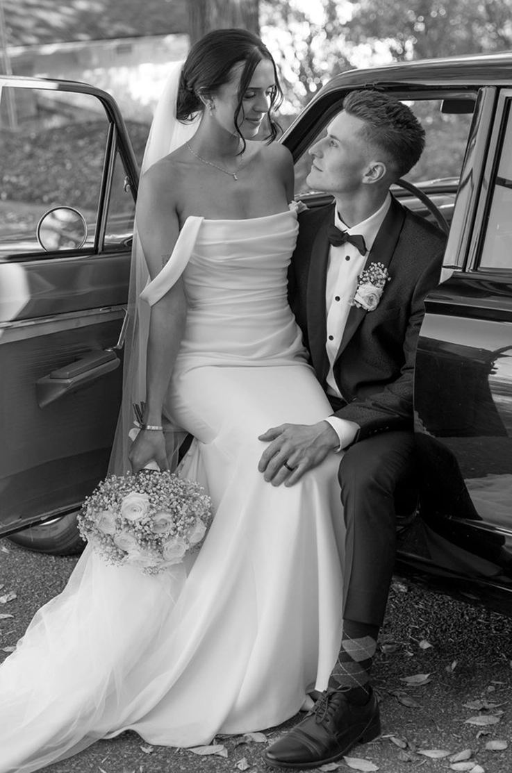 Bride in an off-shoulder fitted wedding dress with a draped bodice and sleeves, sits on groom's lap in a vintage car.