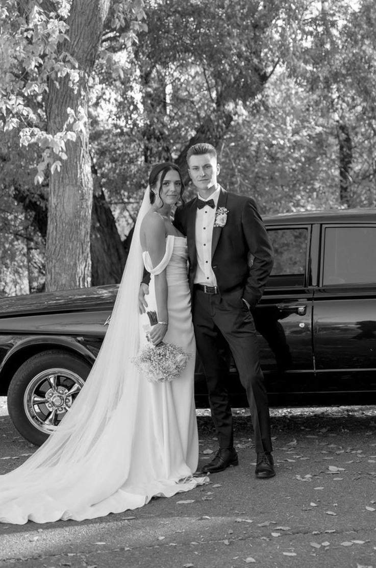 Bride in a draped crepe bodice gown with sleeves and a veil, poses beside her groom in a tux by a car, in black and white. 