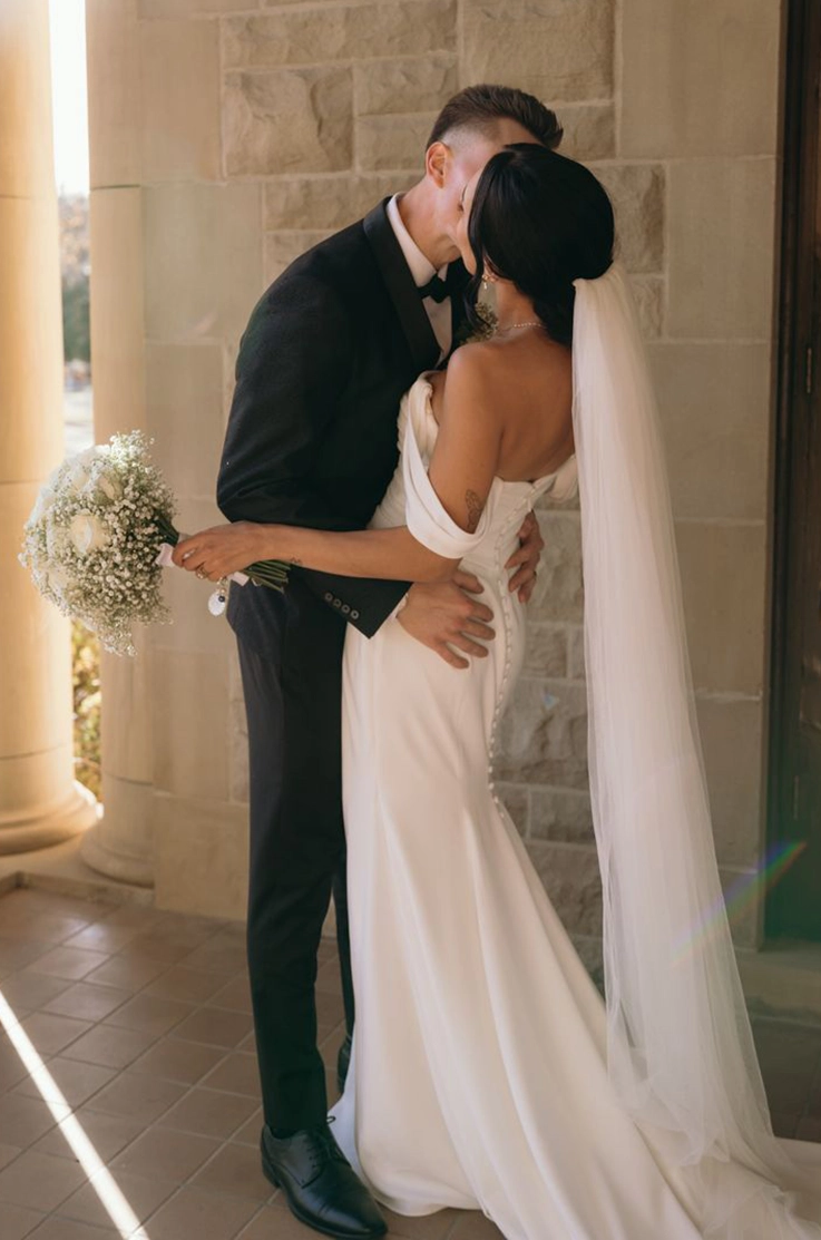 Groom holds his bride in an embrace in a crepe fit and flare gown with a buttoned back, sleeves and a veil at a stone building.