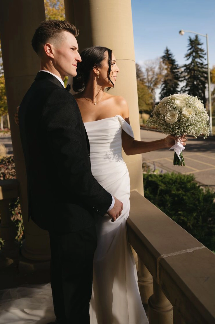 Bride in a draped bodice gown with off-shoulder sleeves, stands by a railing next to her groom outdoors and holds a bouquet.