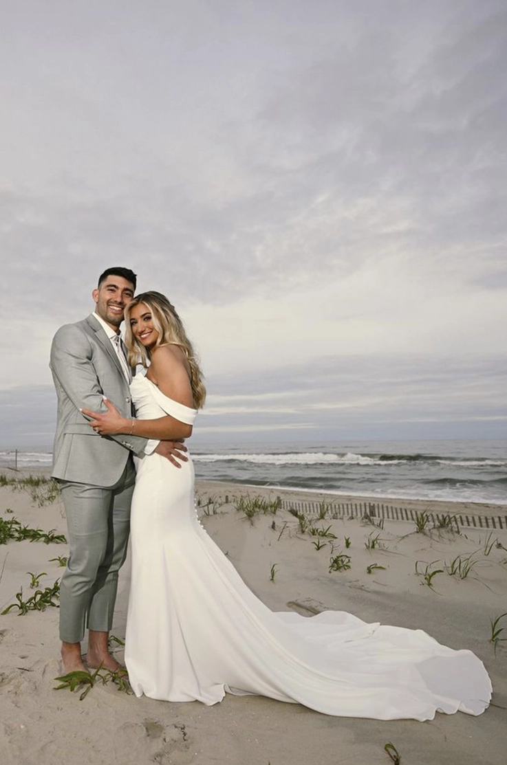 Bride in a crepe fit and flare gown with a draped bodice, sleeves and buttons poses with her groom on a beach shore.