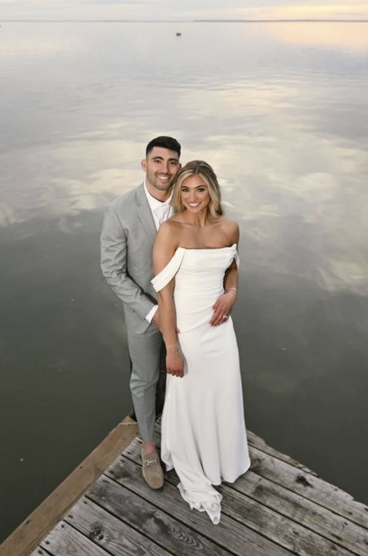 Groom embraces his bride in a draped crepe wedding dress with sleeves, standing on a wooden dock at sunset.