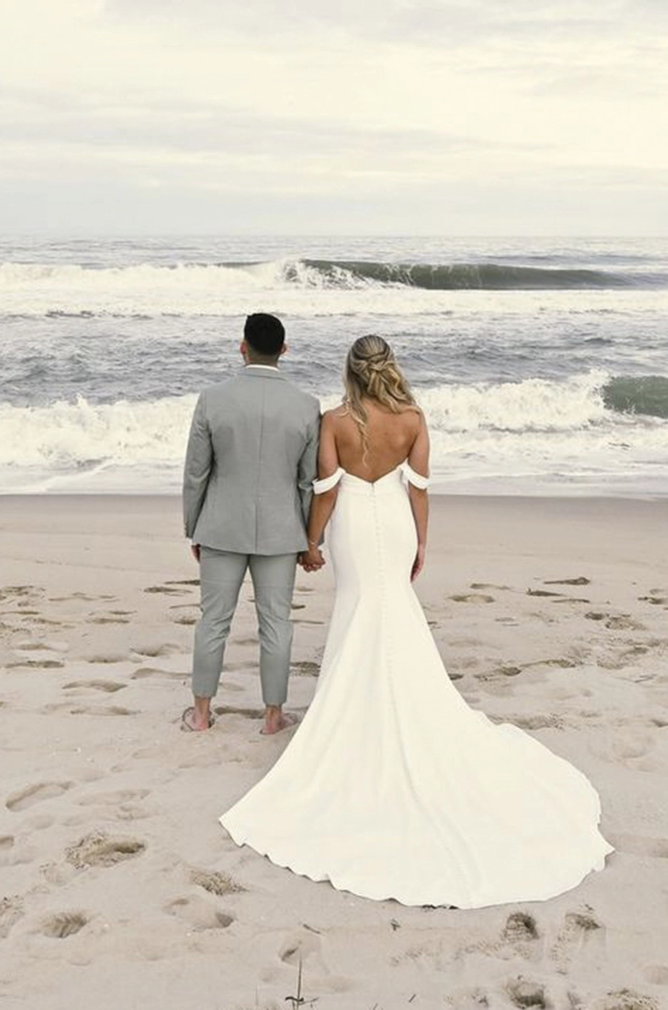 Back of bride in a low back crepe gown with buttons, fit and flare skirt and sleeves, looks out at an ocean next to the groom. 
