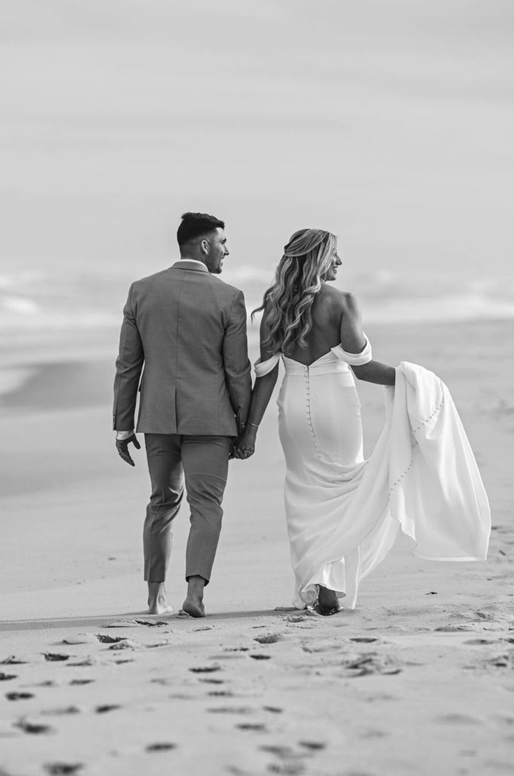 Bride picks up her train in an off-shoulder fit and flare gown with buttons, is walking alongside her groom on a beach.