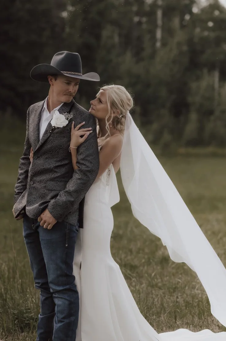 Bride in a fit and flare crepe gown with a flowing veil, holds her groom in an embrace in a cowboy hat, in an open field. 