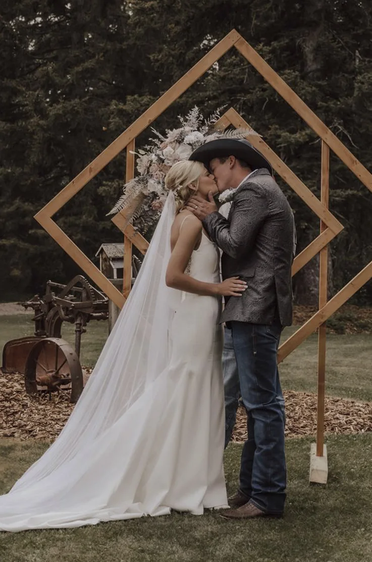 Bride in a spaghetti strap crepe wedding dress with a veil, kisses the groom at a geometric floral arch outdoors.