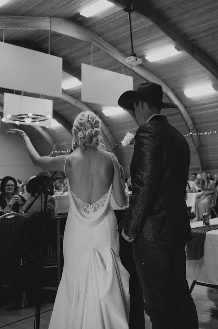 Bride in a fit and flare crepe gown with a low open back and lace detail, stands next to groom in a cowboy hat, in black and white