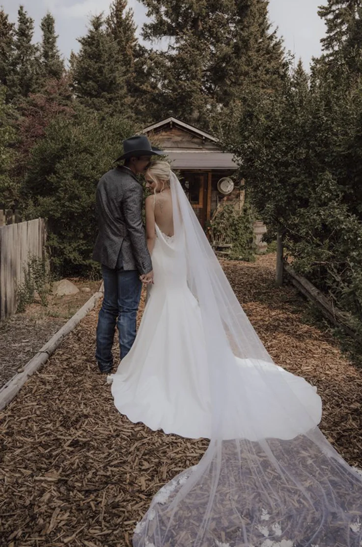 Bride in a fit and flare crepe gown with a scoop neckline and a long veil, poses beside the groom at a forest cabin. 