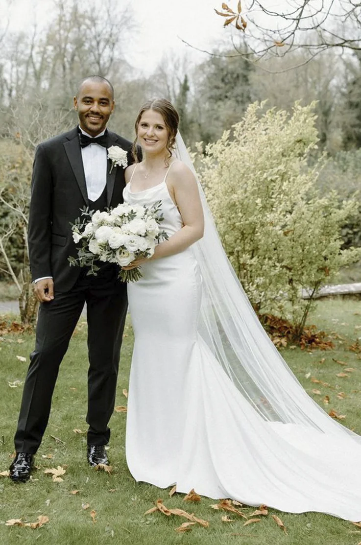 Bride in a cowl neck satin wedding dress with fit and flare skirt and a veil, poses next to her groom outdoors.
