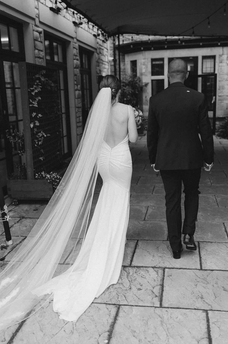 Back of bride in a low back fit and flare gown with a long veil, walks beside her groom towards the venue, in black and white.
