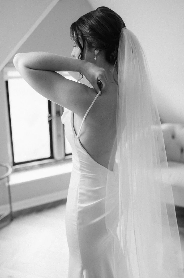 Bride poses in a draped cowl neck satin gown with a fit and flare skirt, holding a white rose bouquet indoors.