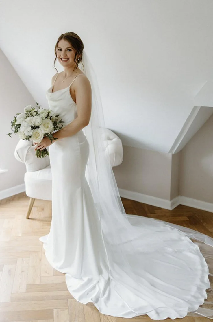 Bride in a charmeuse satin fitted gown with spaghetti straps and a veil, poses by bright windows indoors, in black and white. 