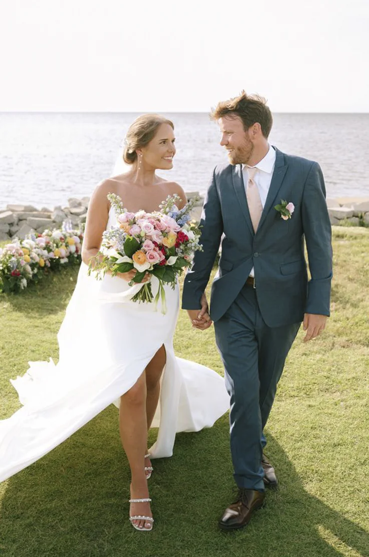 Bride in a fit and flare crepe wedding dress with a skirt slit and veil, is walking along a waterfront beside her groom.