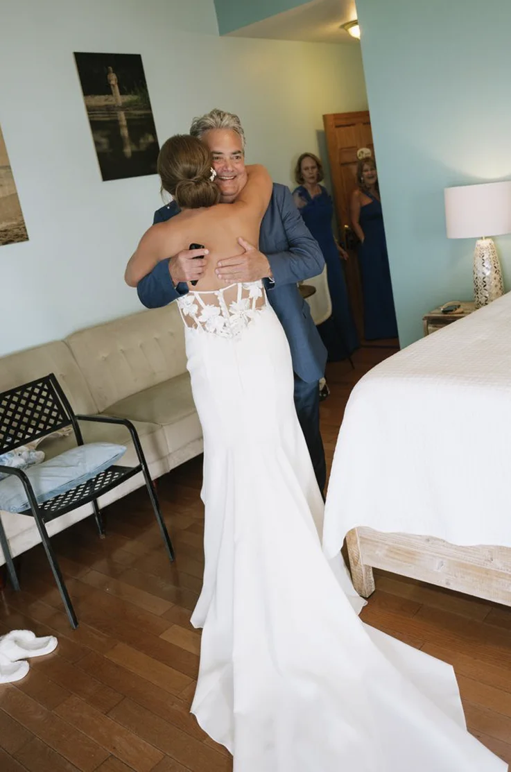 Bride is in a crepe fitted gown with a sheer corset back, hugs her father in a bridal suite.