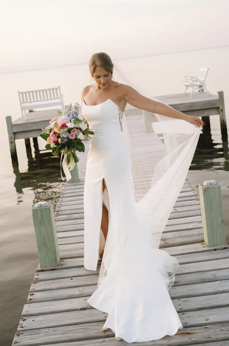 Bride in a scoop neck crepe gown with a skirt slit, poses and holds her veil on a lakeside dock.