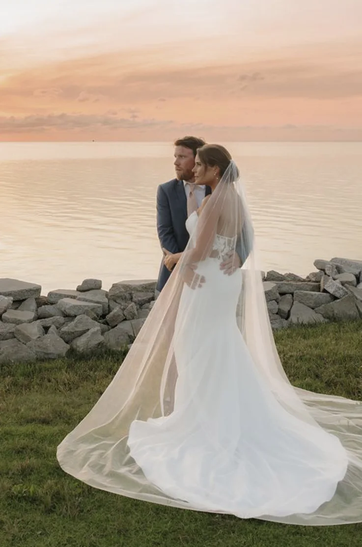  Bride in a strapless crepe gown with a sheer back and flowing veil, poses with her groom by a waterfront at sunset.