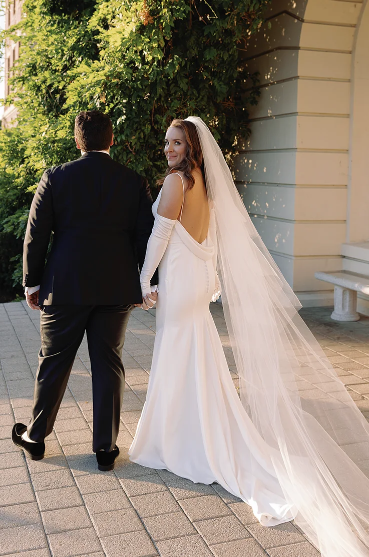 Back of bride in a draped cowl charmeuse satin gown with sleeves, and a veil, walks alongside her groom outside venue. 