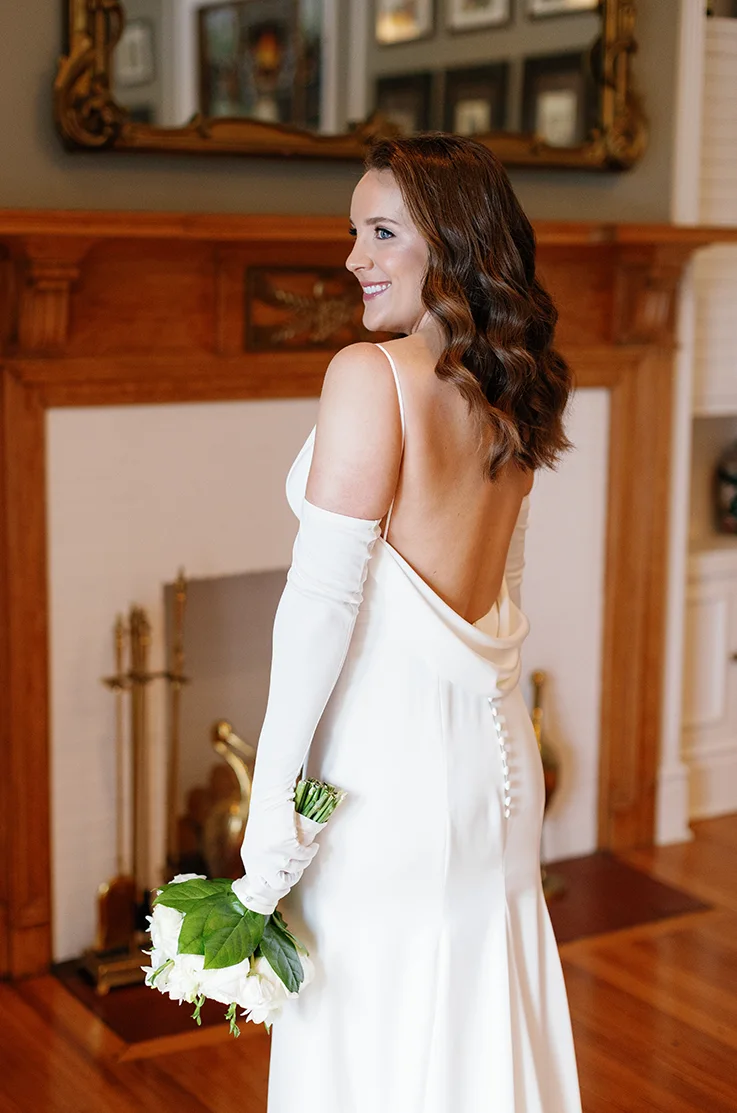 Bride poses in a fitted charmeuse satin wedding dress with a low back and sleeves, by a fireplace and holds a bouquet.
