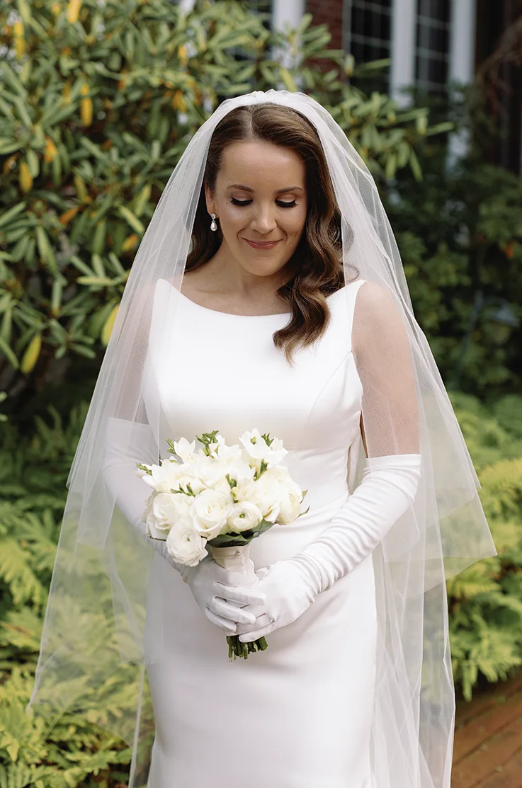 Close-up of bride in a sleeveless charmeuse satin gown with a bateau neck, sleeves and a veil, holds a bouquet outdoors. 
