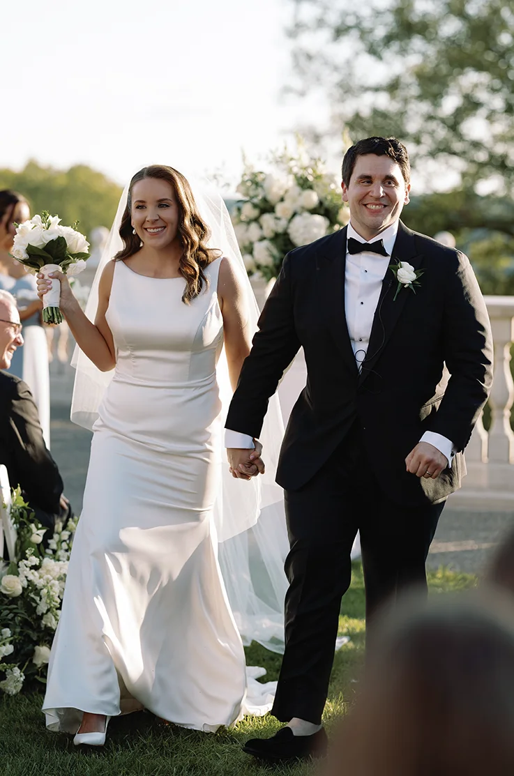 Bride in a sleeveless fit and flare gown with a bateau neckline and veil, walks hand in hand with her groom down the aisle.