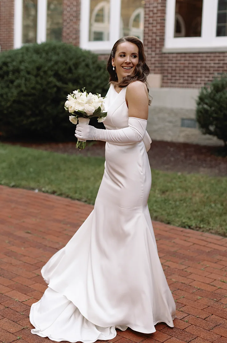Bride in charmeuse satin fit and flare gown with a bateau neck and sleeves, is holding a bouquet on a brick path.