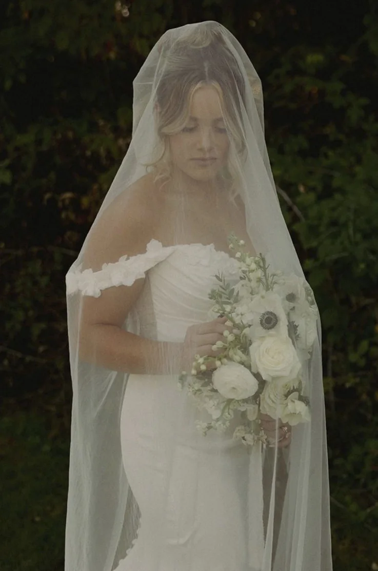 Bride in a satin fit and flare wedding dress with lace detail, poses under a veil and holds a bouquet in dramatic lighting.