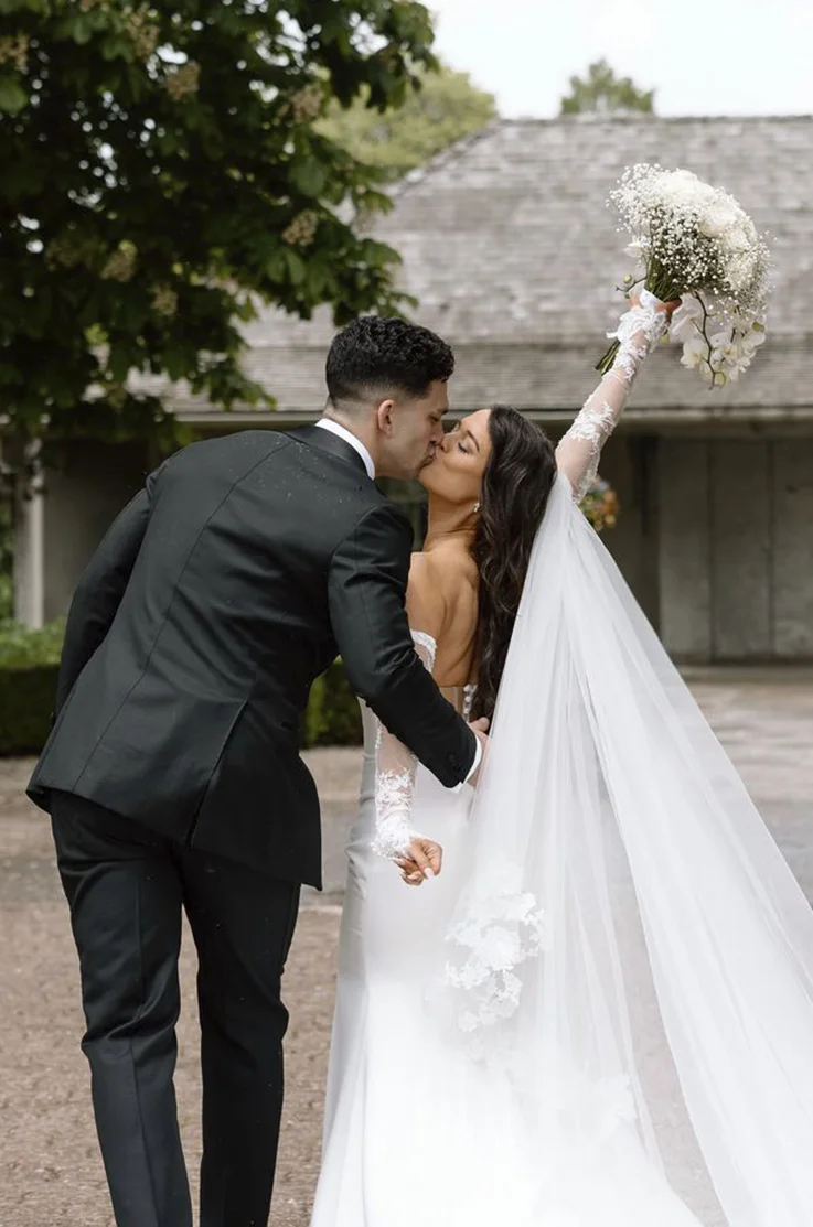 Groom kisses bride in a crepe gown with sheer sleeves and a lace-trim veil, holding up a bouquet at a garden estate.