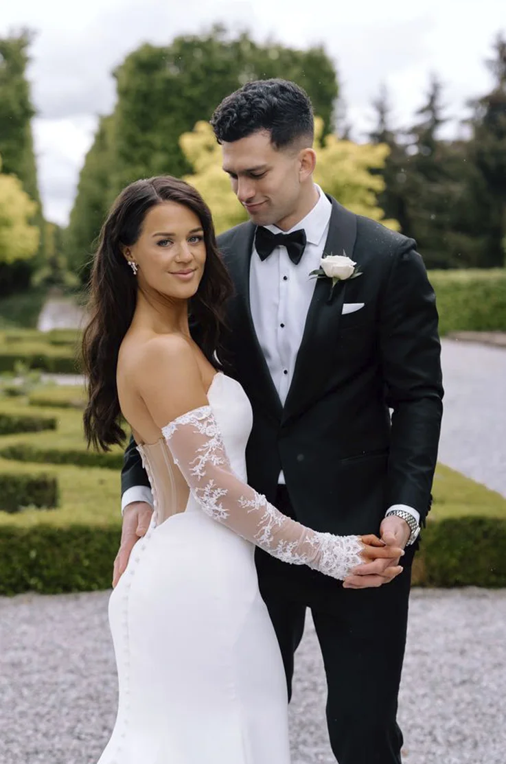 Bride poses in a strapless crepe wedding dress with a tulle sheer back and sleeves, with her groom in a courtyard garden.