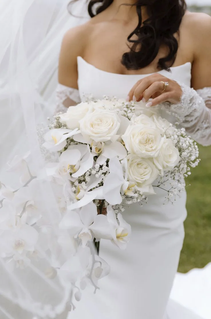 Close-up of bride in a crepe fitted gown with a scoop neck, sheer lace sleeves and a flowing veil, holding a rose bouquet. 