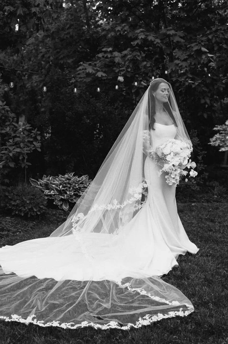Bride in a scoop neck crepe gown with lace and a long veil, is holding a large bouquet outdoors in black and white. 