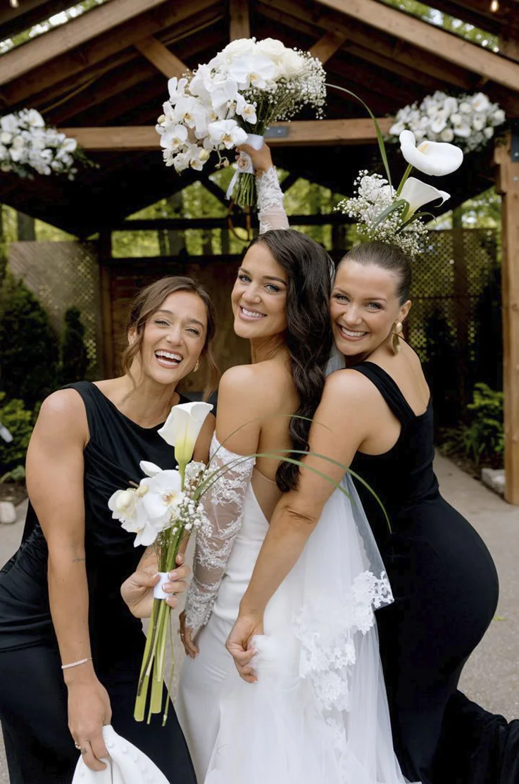 Bride looks over her shoulder in a crepe strapless gown with sleeves and a veil, between bridesmaids in black dresses. 
