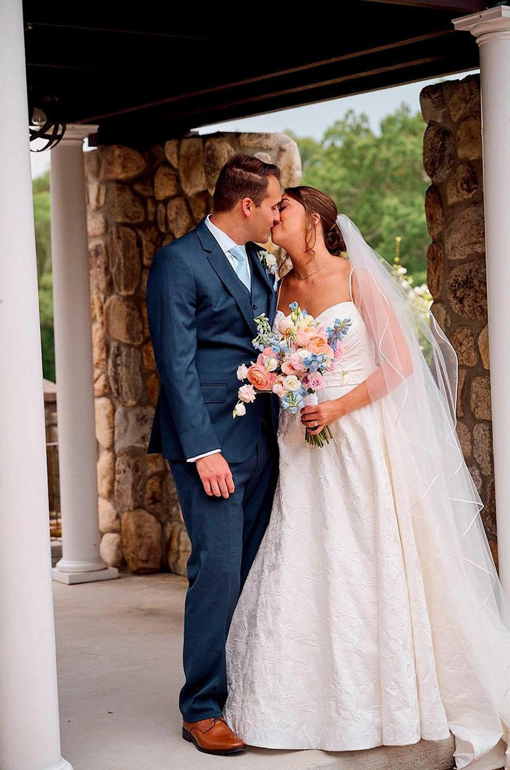 Groom kisses his bride in a sleeveless brocade ballgown with a flowing veil, against a tall white pillar on a stone terrace.