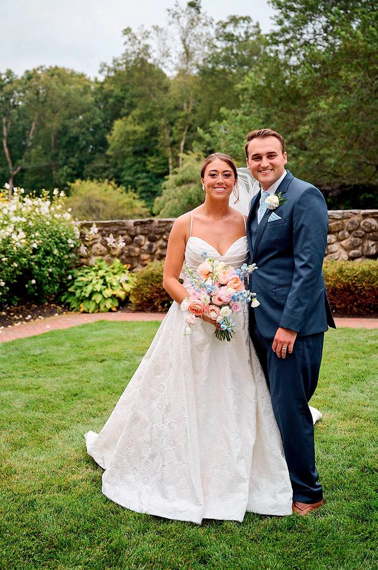 Bride in a sweetheart neck brocade wedding dress, is holding a colorful bouquet beside her groom in a garden.