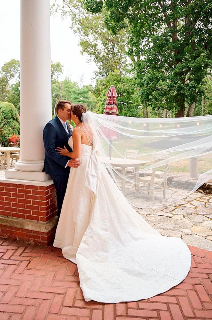 Groom kisses his bride in a sleeveless brocade ballgown with a flowing veil, against a tall white pillar on a stone terrace.