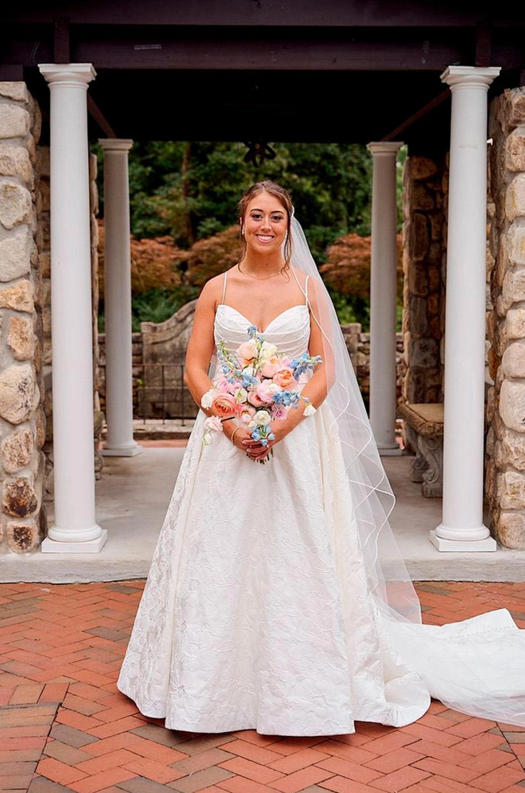 Bride in a sweetheart neck ballgown with a veil, kisses her groom between stone pillars, holding a pastel bouquet.