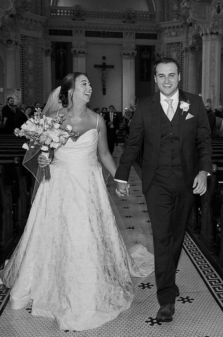 Bride in a sweetheart neck floral print gown with a veil, walks hand in hand with her groom down a Church aisle.