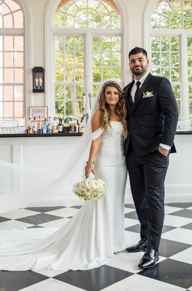 Groom poses beside bride in an off-shoulder draped bodice gown with a flowing veil, in front of a white bar counter.